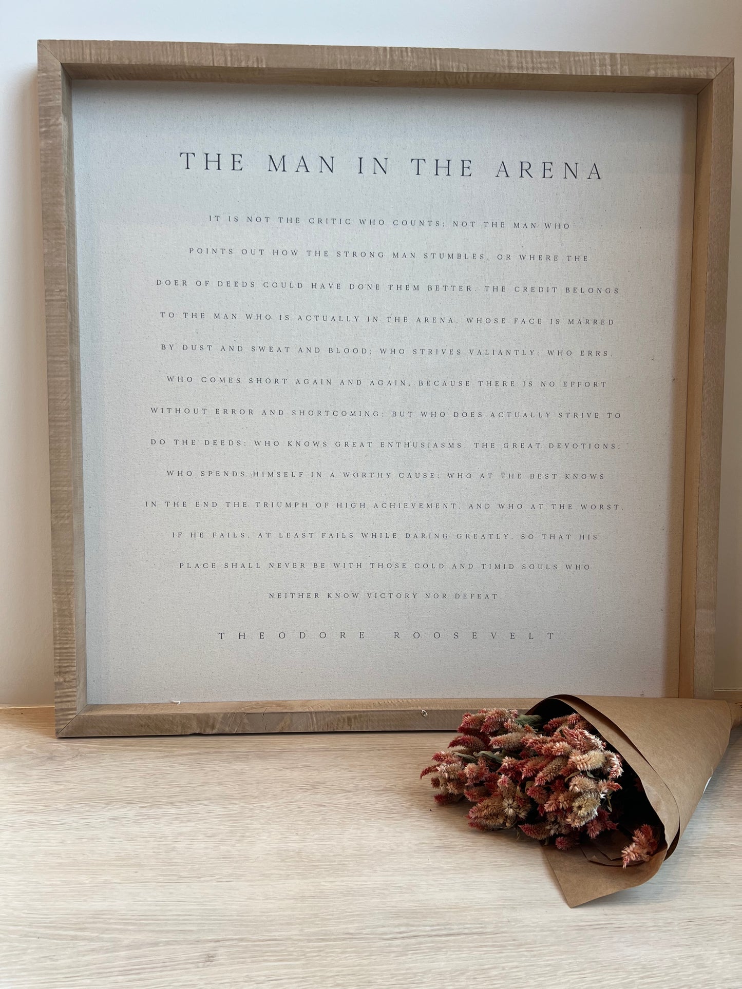 Framed text art with a decorative bouquet of dried flowers on a light wooden surface.
