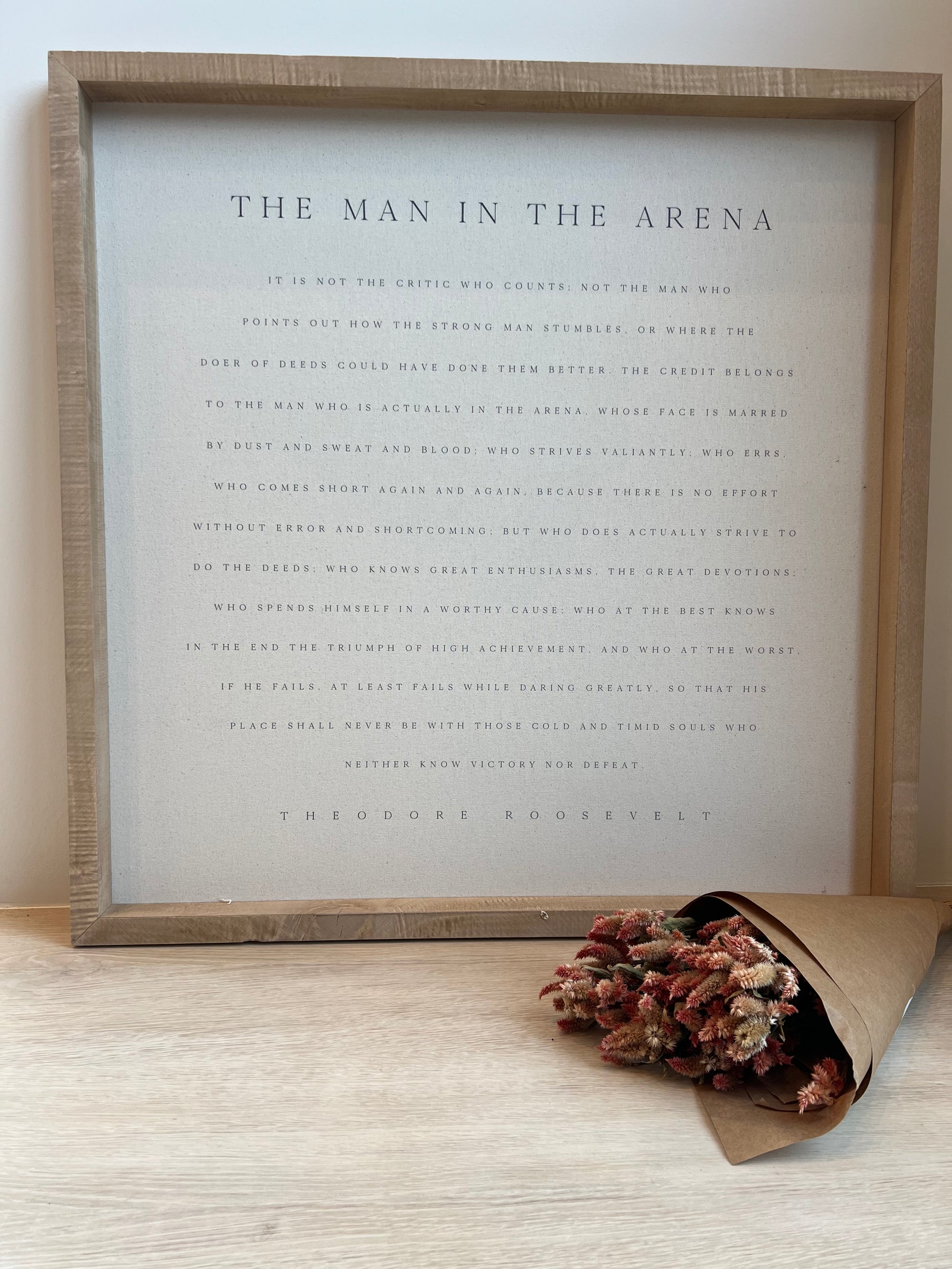 Framed text art with a decorative bouquet of dried flowers on a light wooden surface.