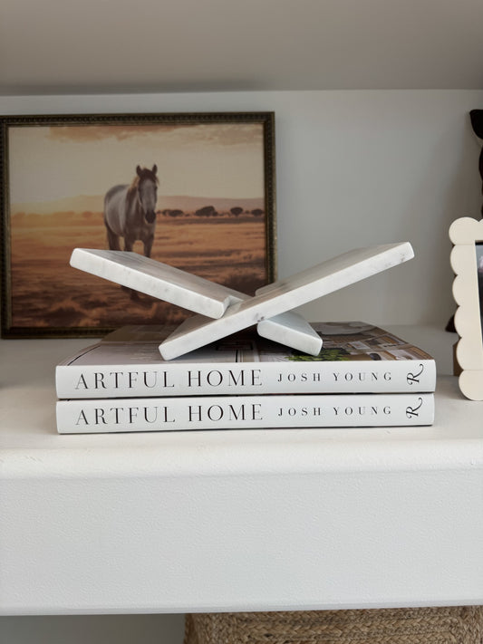 Stack of books titled 'Artful Home' by Josh Young on a shelf with a horse painting in the background.